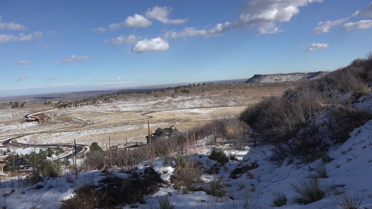 One second into the clip, the Golden, Colorado view holds on snow patches, dry winter brush, a curved road, low buildings, distant ridgeline, and no visible people.