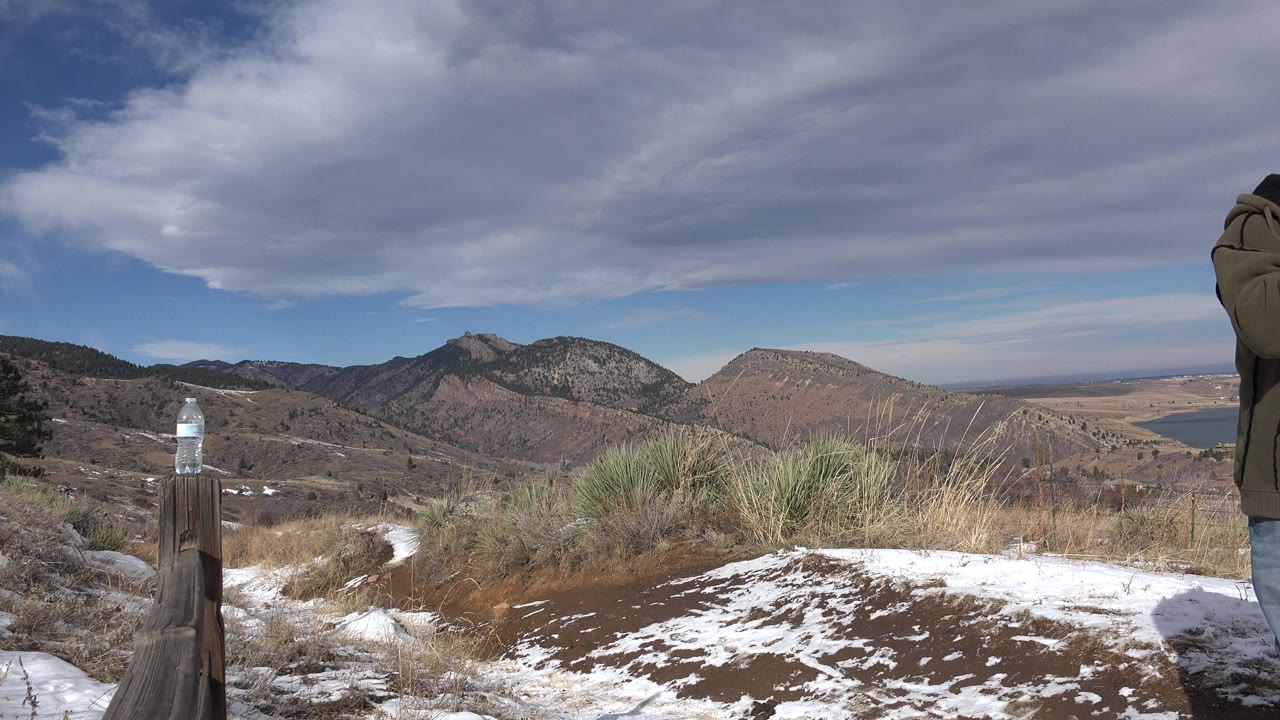 Mountain overlook with snow patches, dry grasses, a water bottle on a post, and a partial person at right.