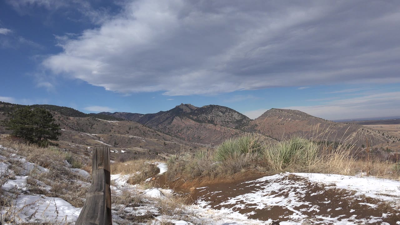 Opening view of a Golden, Colorado hillside with patchy snow, dry grasses, a wooden post at left, distant mountains, layered clouds, and no visible people.