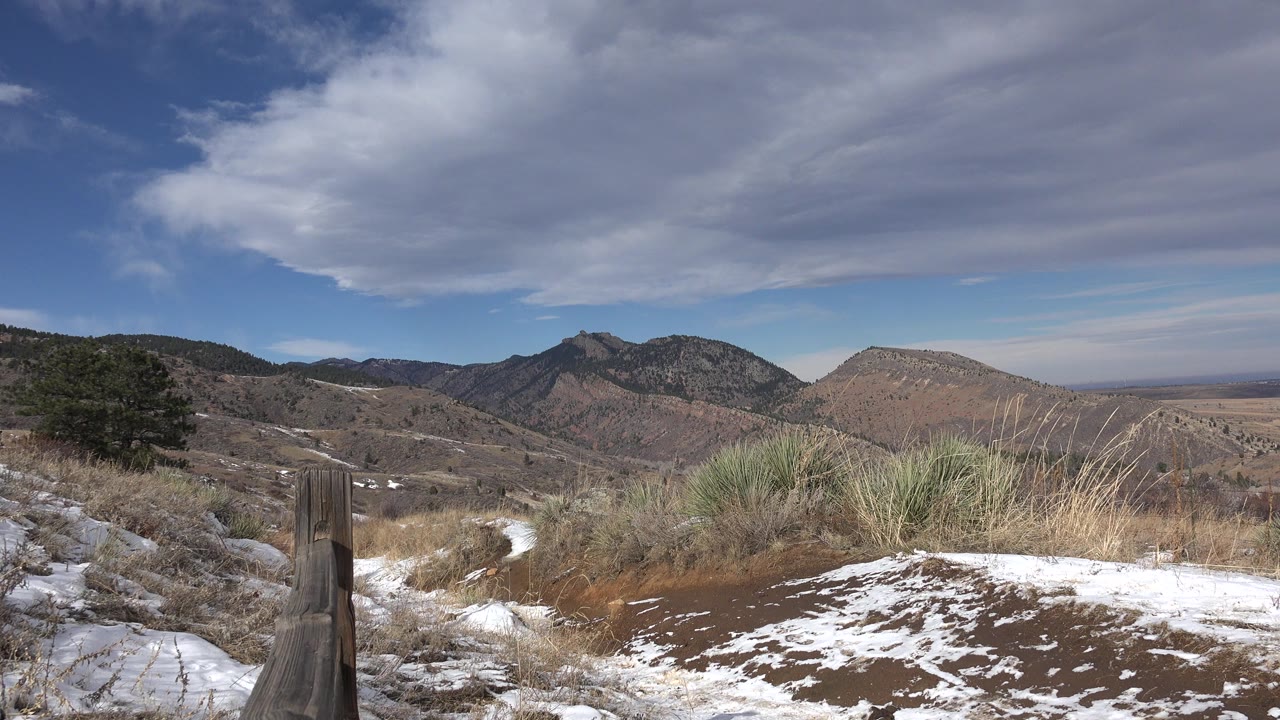 Opening frame of a Golden, Colorado hillside with patches of snow, a wooden fence in the foreground, dry grass, distant mountains, broad clouds, and no visible people.
