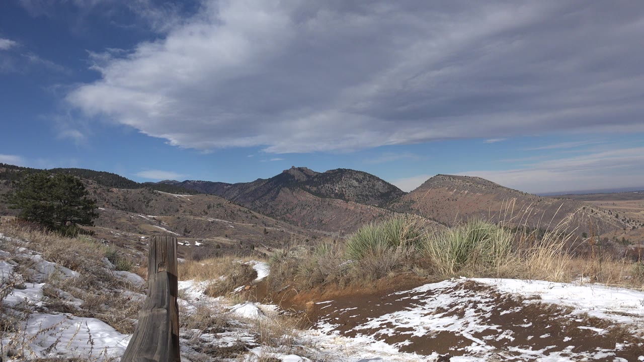 Opening view in Golden, Colorado showing patchy snow, dry grass, a dark trail marker at left, distant mountains, layered clouds, blue sky, and no visible people.