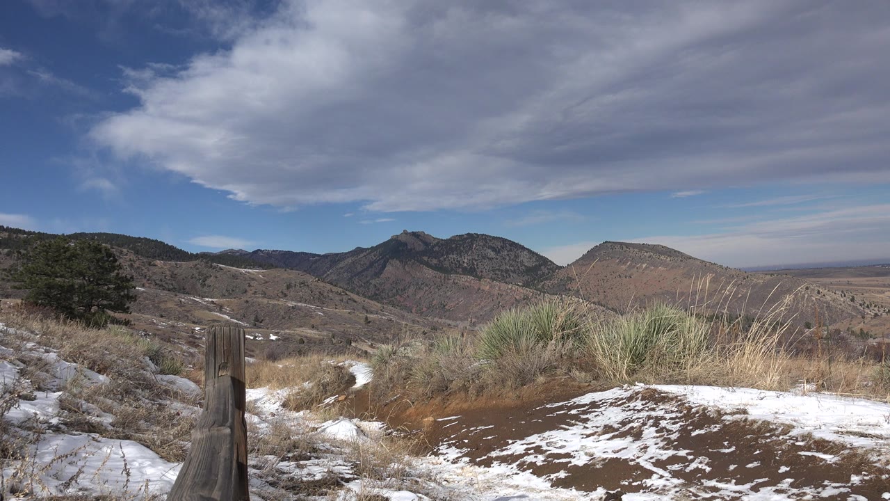 Opening frame of Golden, Colorado foothills with snow patches, dry grasses, a wooden post at left, distant mountains, layered clouds, and no visible people.