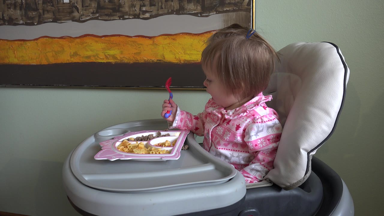 At 0:00 in Westminster, Colorado, a young child sits side-on in a gray high chair, holding a red-and-blue fork above a divided plate of food with abstract wall art behind them.