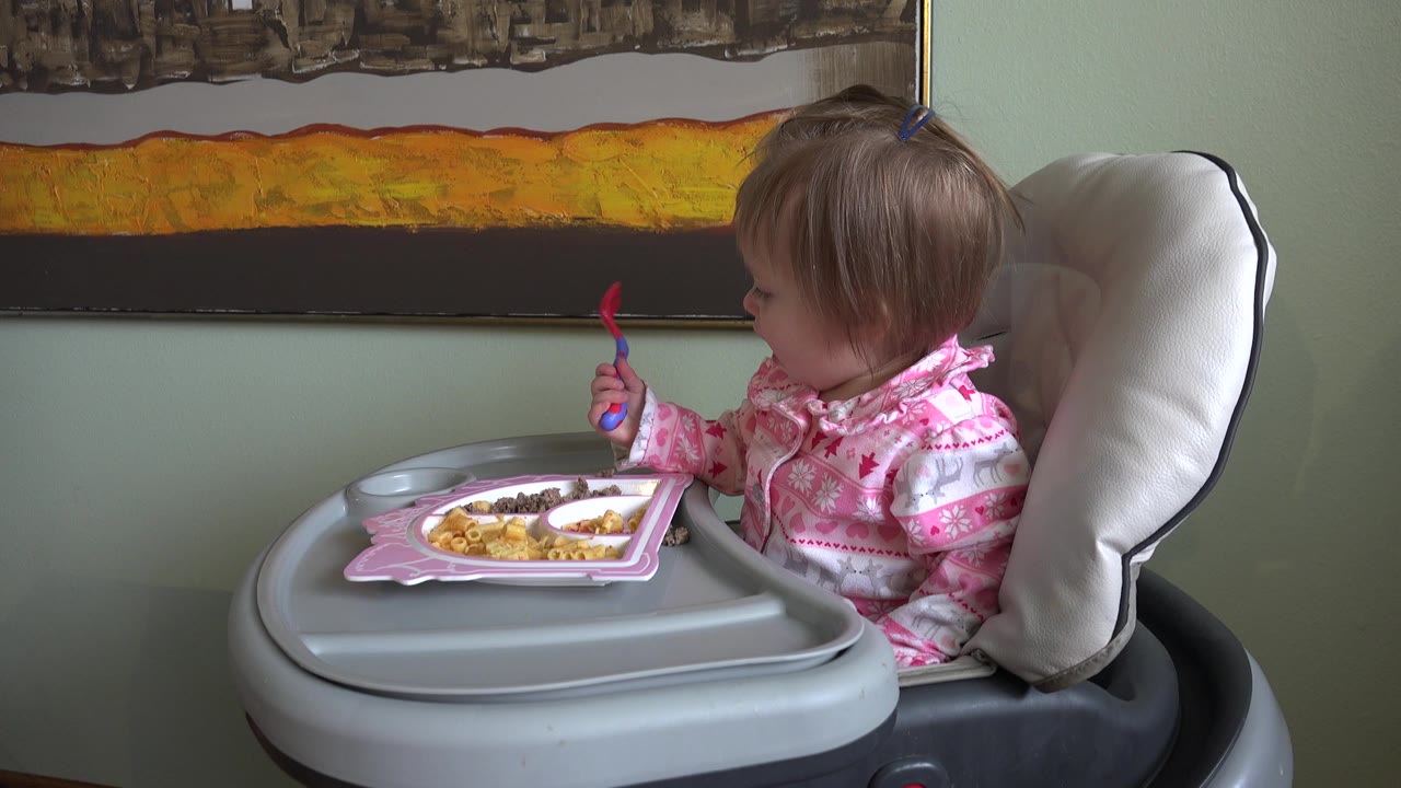At 0:01 in Westminster, Colorado, the child remains turned toward the plate, with the fork raised in one hand and the high-chair tray filling the foreground.