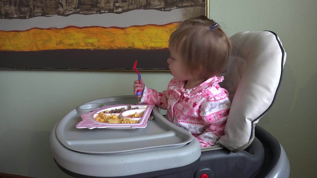 At 0:02 in Westminster, Colorado, the child looks farther to the right from the high chair, with the fork still raised and the plate of food on the tray below.
