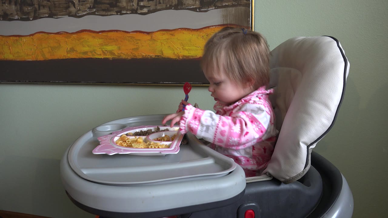 At 0:00 in Westminster, Colorado, a baby girl in pink pajamas sits in a gray high chair and looks down toward a plate of food beneath a large framed artwork.
