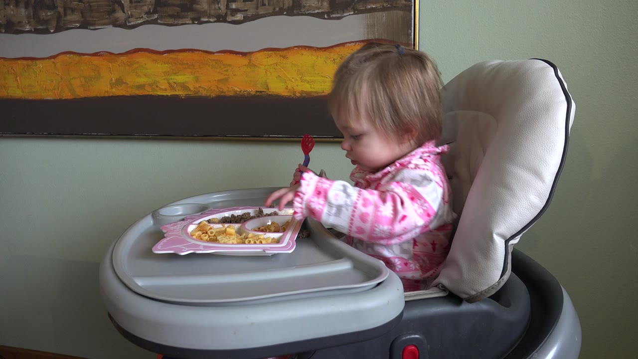 At 0:01 in Westminster, Colorado, the baby remains turned toward the high-chair tray, holding a red spoon beside a pink plate of food with the framed artwork above her.