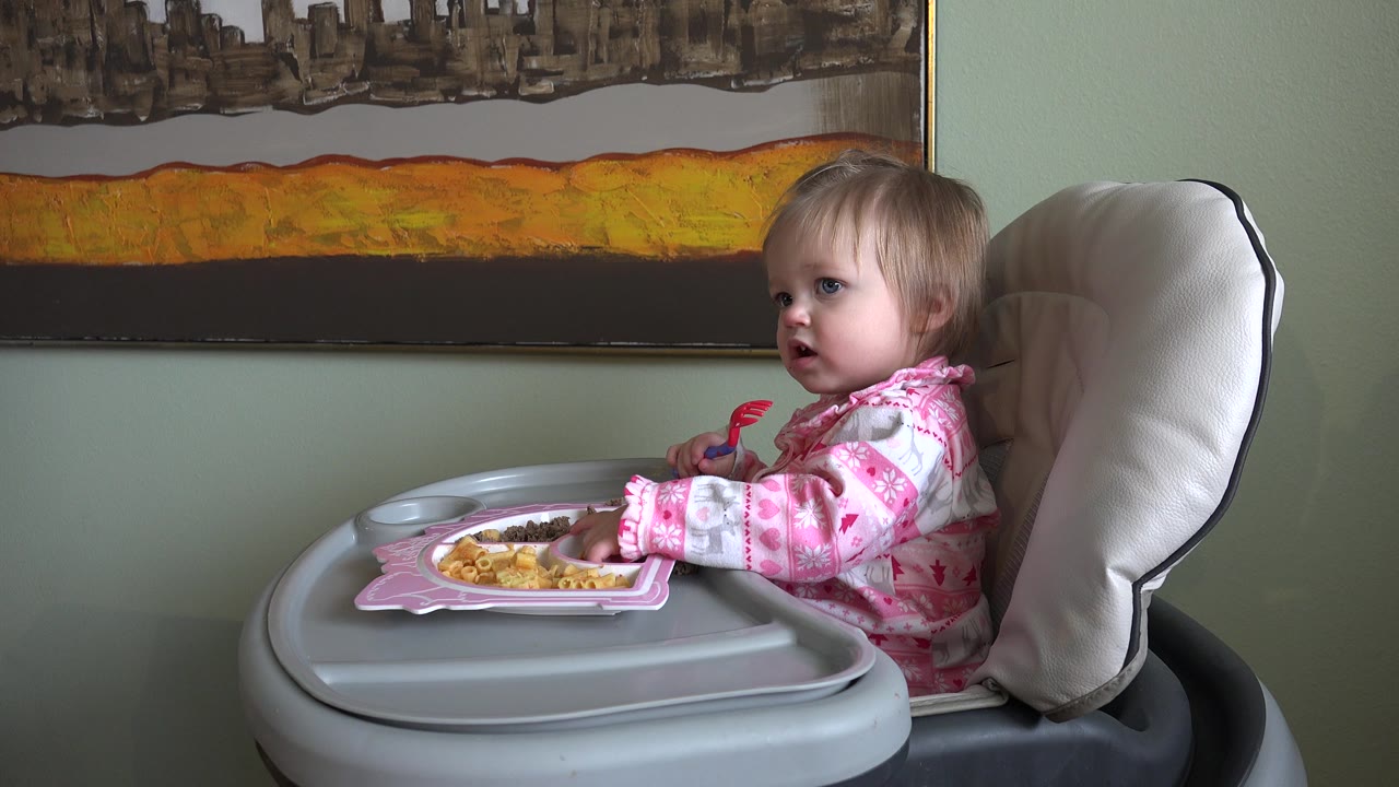 At 0:00, a toddler girl in pink pajamas sits in a gray booster seat in a Westminster, Colorado home interior, facing left with food on a tray and abstract wall art behind her.
