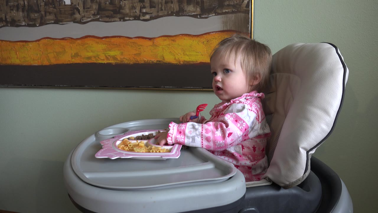 At 0:02, the toddler sits upright with her mouth slightly open, the divided meal plate centered on the tray and the large abstract artwork spanning the wall above.
