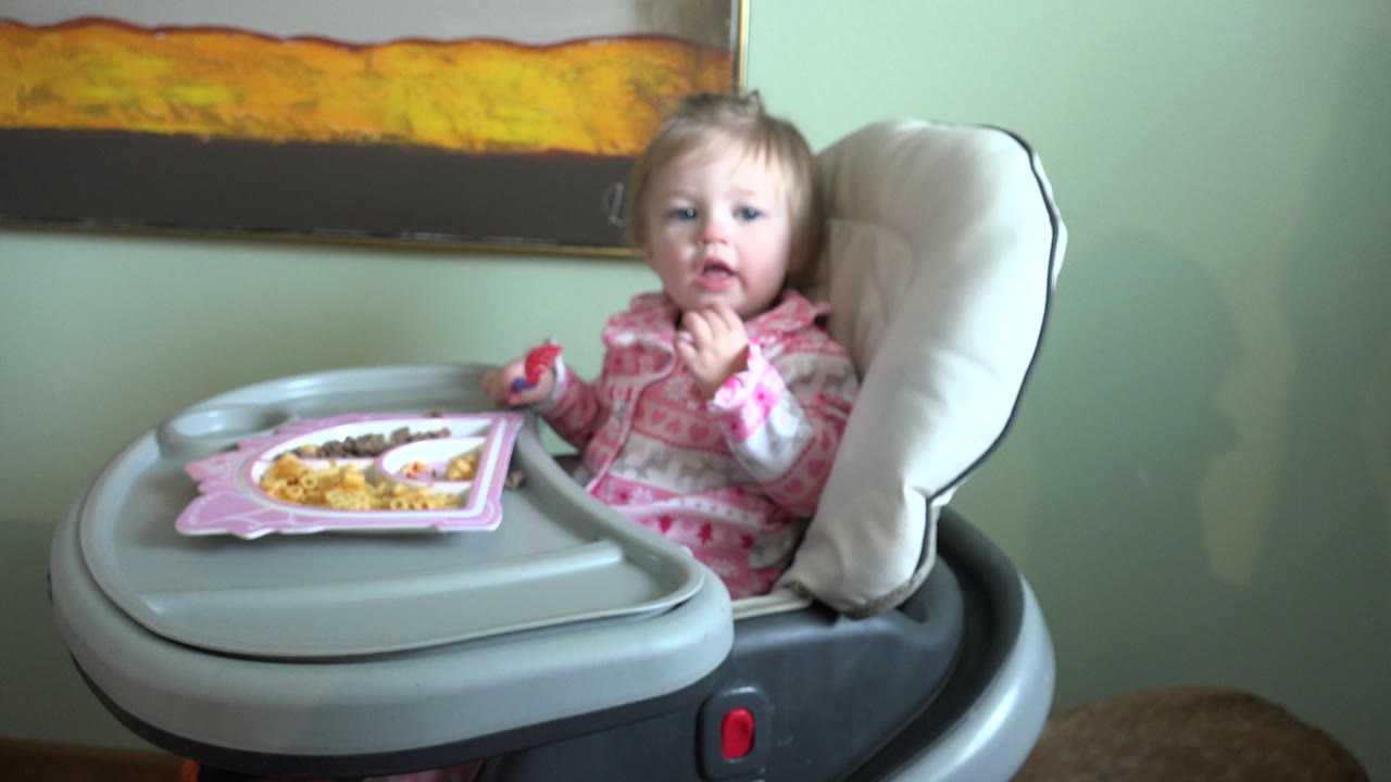 A young child sits in a gray highchair in Westminster, Colorado, with a pink tray of food, pale green wall, white cushion, and framed artwork behind them.