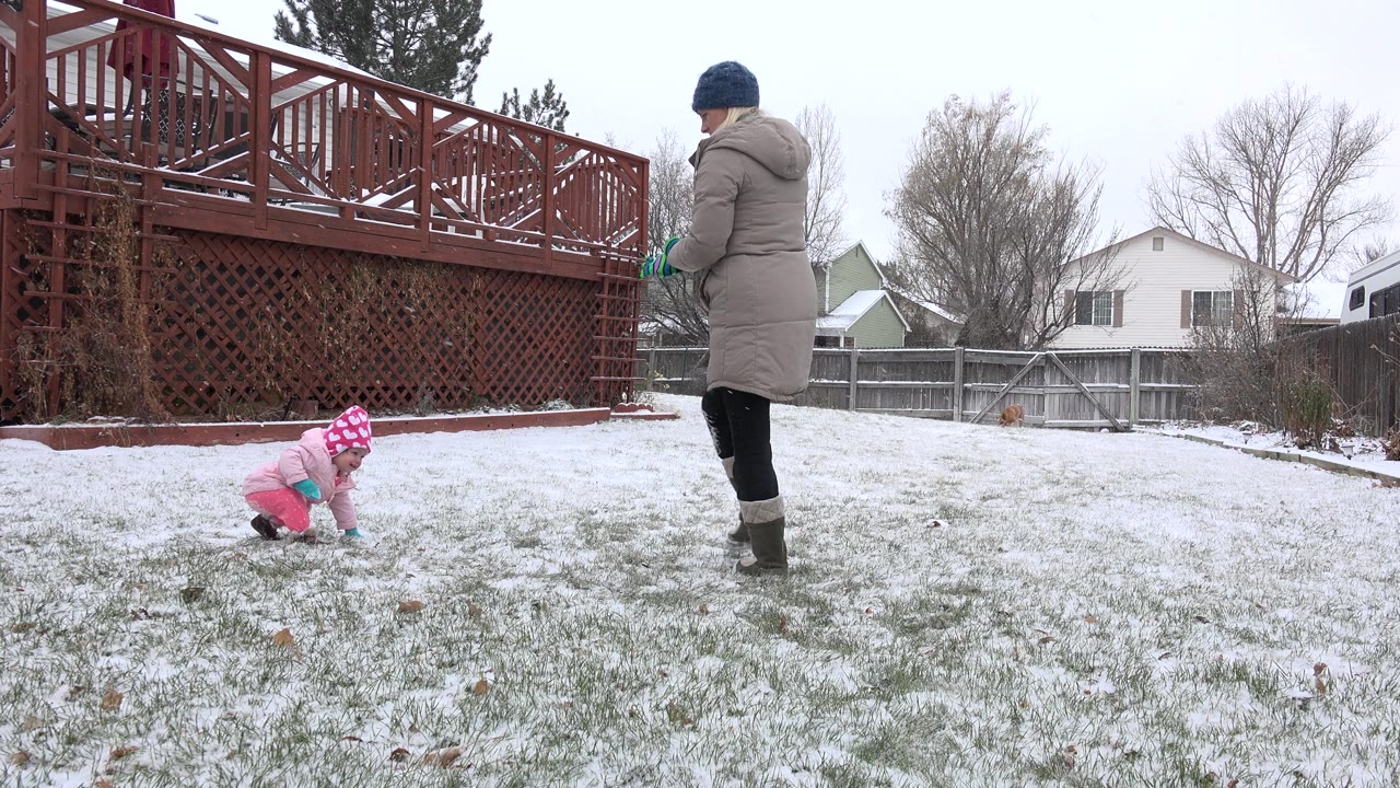 In Westminster, Colorado, a bundled toddler crawls on a thin layer of backyard snow while an adult stands near a red wooden deck and fence.