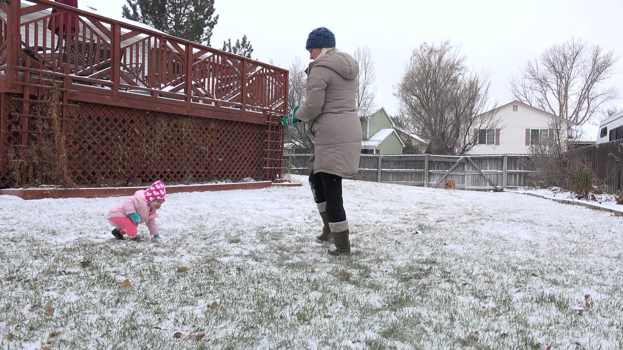A wide Westminster backyard frame shows the adult beside the deck while the toddler continues crawling across patchy snow toward the open lawn.