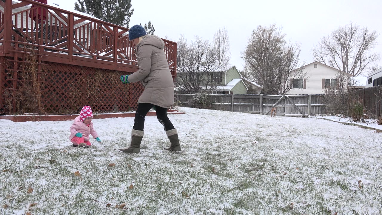 Adult walks toward a toddler sitting on snowy grass in the backyard.