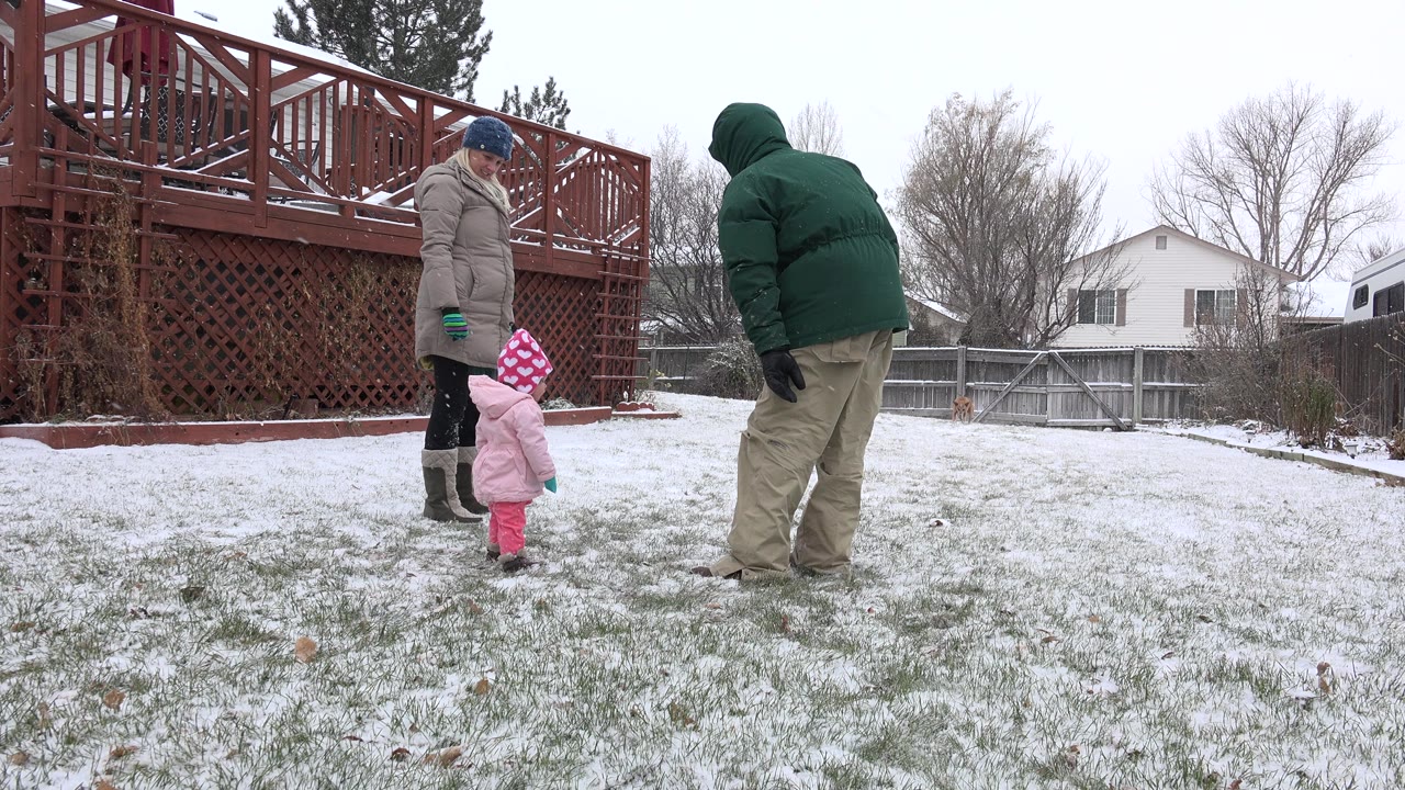 Two bundled adults and a toddler stand in a snowy backyard near a red deck.