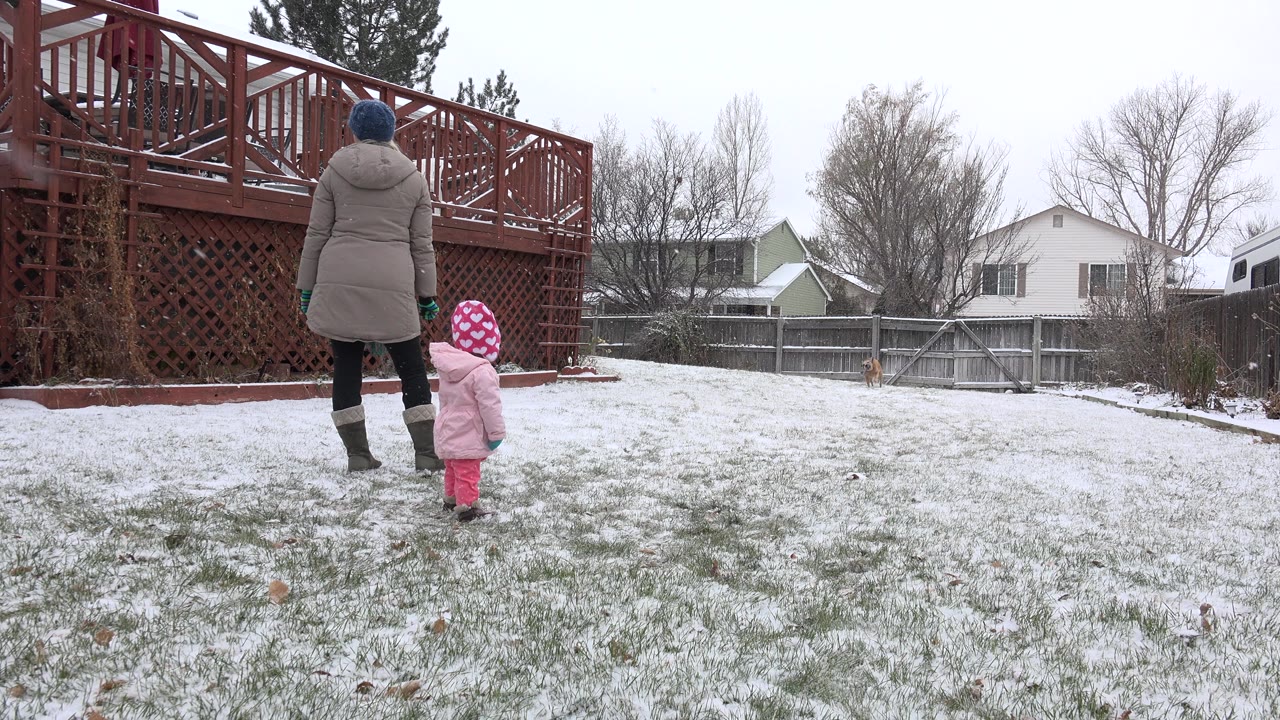 In Westminster, Colorado, a woman in a gray coat and a small child in pink winter clothing stand on a lightly snow-covered backyard lawn near a red wooden deck.