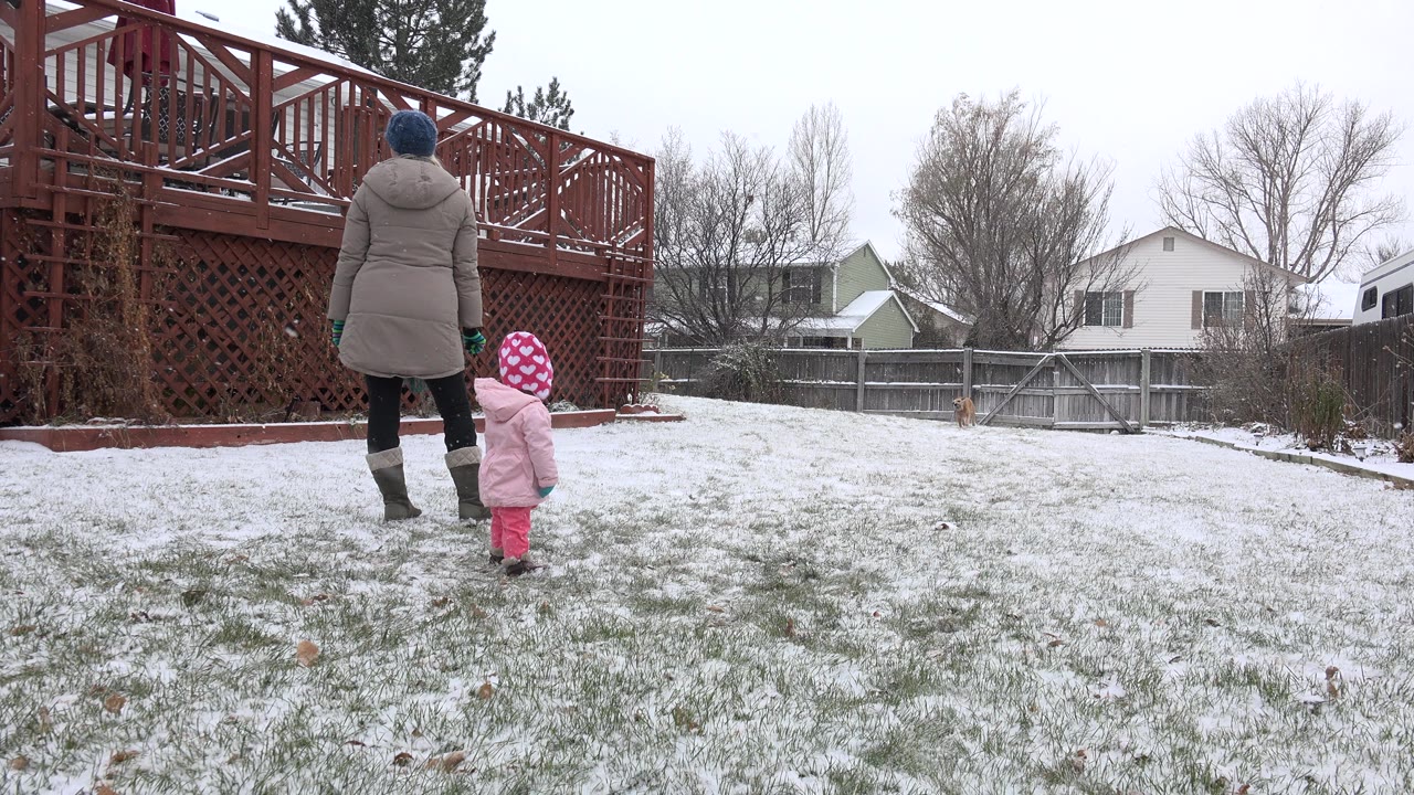 The woman and child remain near the left side of the snowy Westminster backyard, facing toward the open yard with fences and houses behind them.