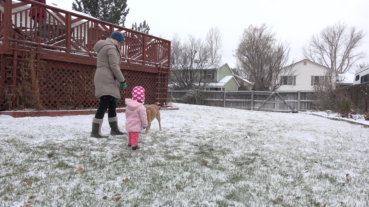 In Westminster, Colorado, a bundled adult and small child stand near a tan dog on a lightly snow-covered backyard lawn beside a red wooden deck.