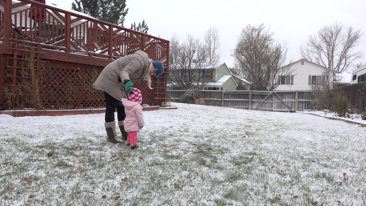 In Westminster, Colorado, an adult in a winter coat bends toward a small child in pink snow clothes on a lightly snow-covered backyard lawn, with a red deck and neighboring houses behind them.