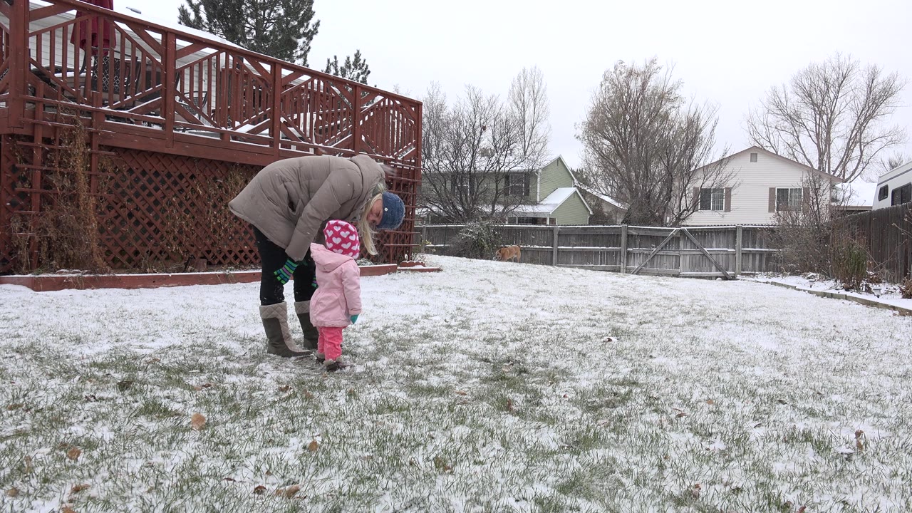 Adult bends toward child while both stand on a snowy backyard lawn.