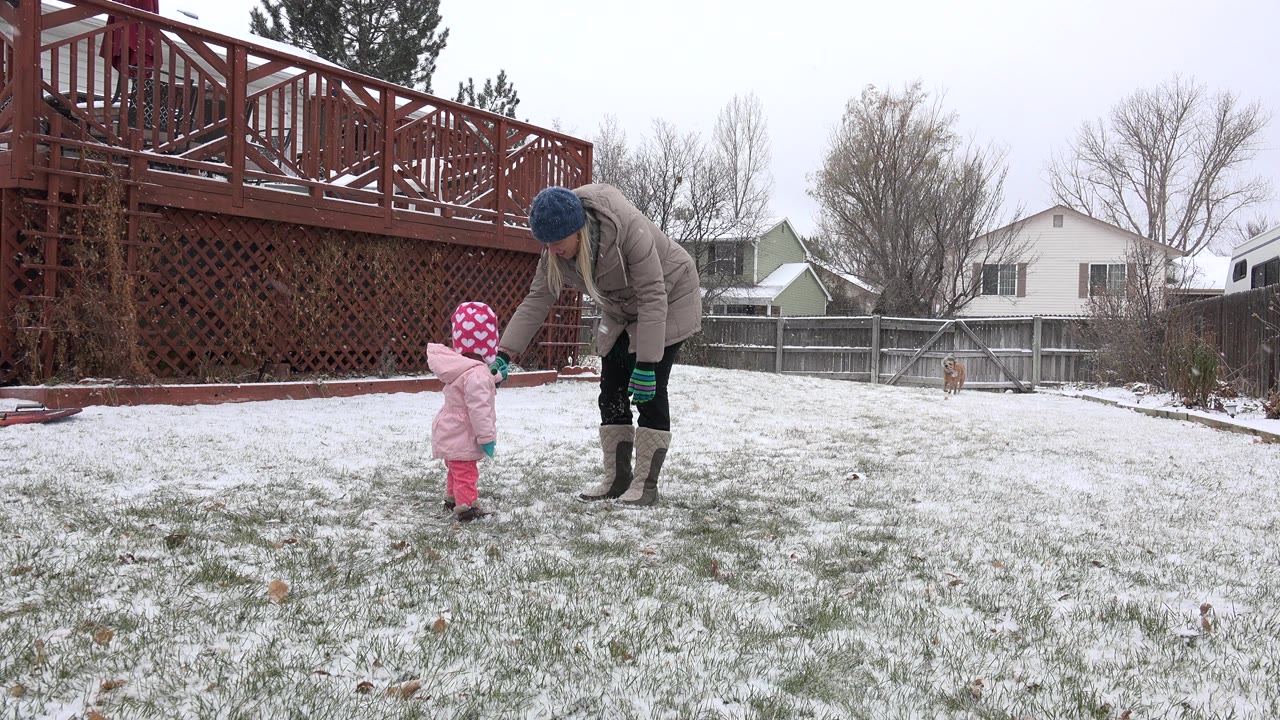 Woman bends toward a small child on a snowy backyard lawn.