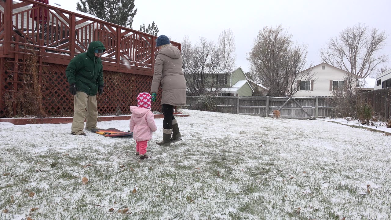 In Westminster, Colorado, two bundled adults stand near a sled or mat while a toddler in pink winter clothes faces them on a lightly snow-covered backyard lawn.