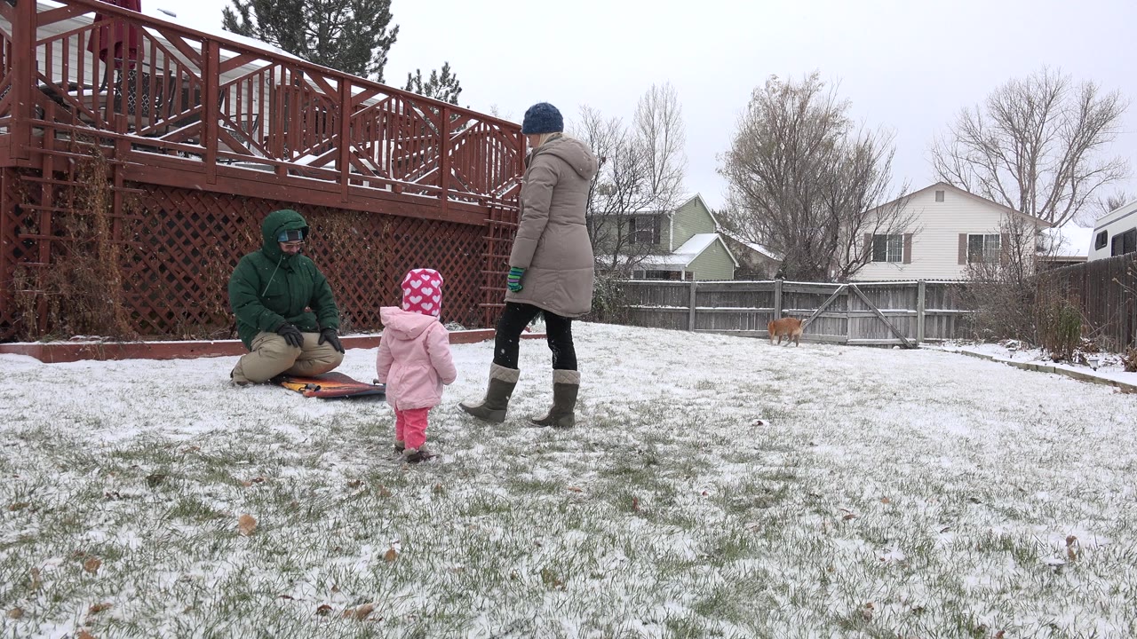 Adult kneels by a sled or mat as toddler and another adult stand in the snow.