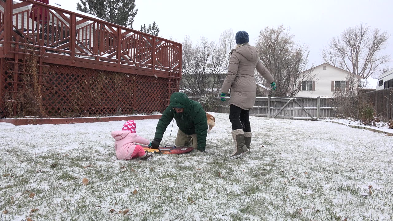 Baby in pink winter clothes near a sled with two adults in a snowy backyard.