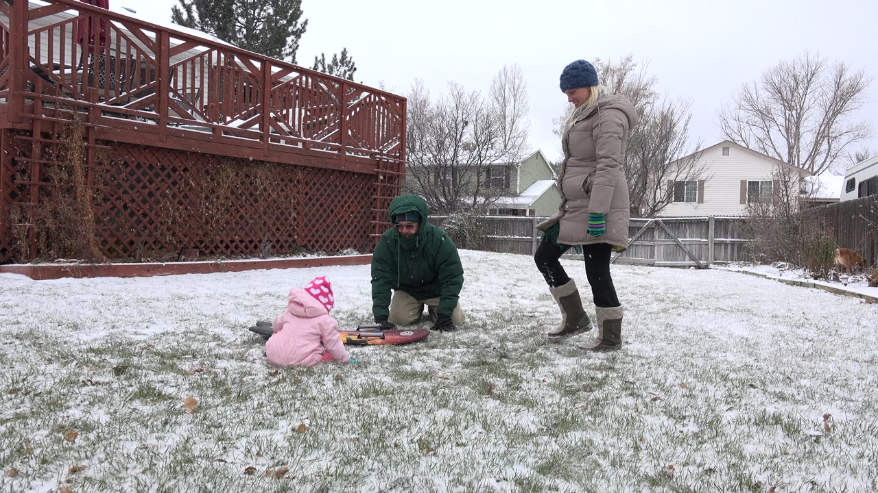 Child in pink winter clothes sits near a sled as two adults stand and kneel in a snowy yard.