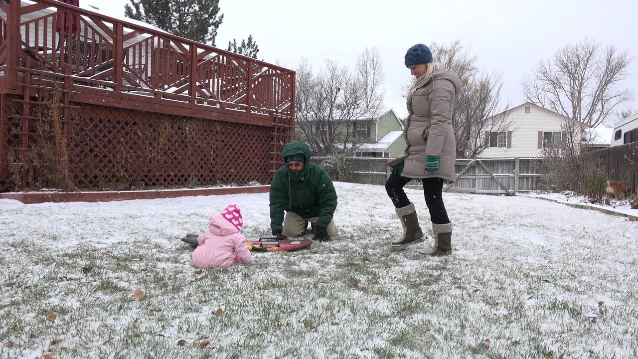 In Westminster, Colorado, the child remains seated on the snowy grass facing the kneeling adult, with the standing adult slightly to the right and homes and fencing behind them.
