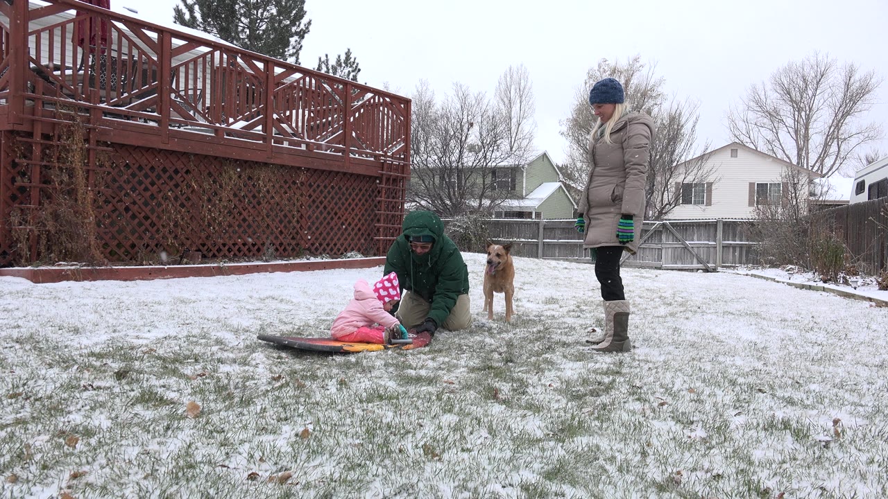 Child on a sled with adults and a dog in a snowy backyard.
