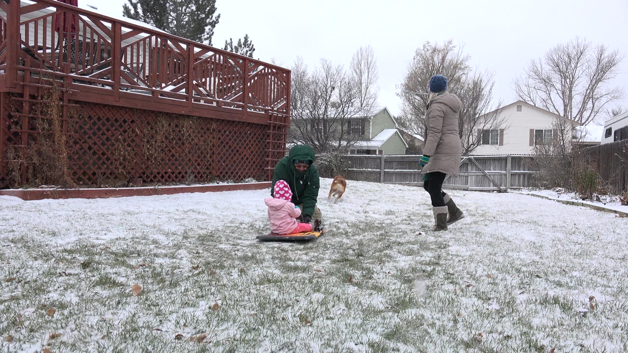 In Westminster, Colorado, the child remains seated on the sled while the crouching adult faces them and a second adult stands to the right on the snowy lawn.
