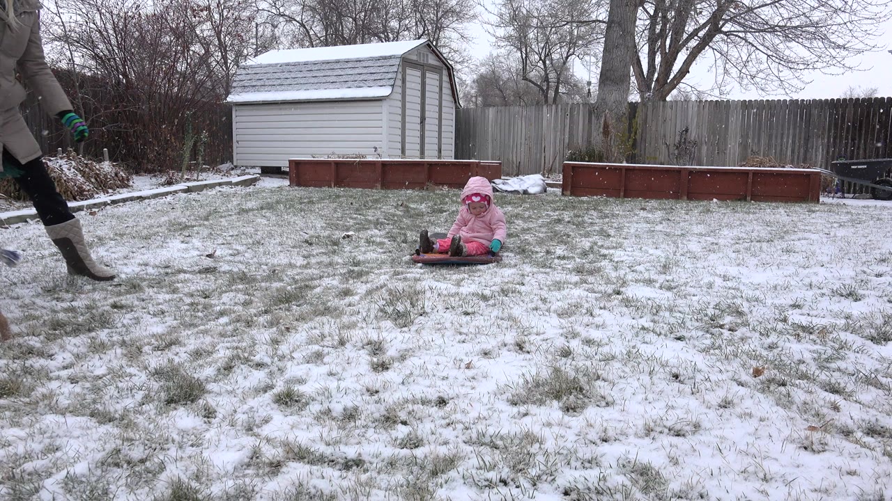 Toddler in pink winter clothing sits on a snowy backyard lawn as an adult leg enters from the left.