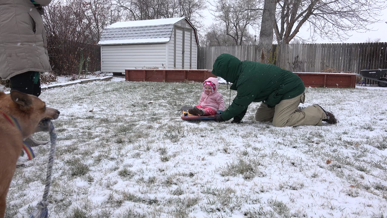 In Westminster, Colorado, a child in pink winter clothing sits on a small sled while an adult in a green coat kneels beside them in a snowy fenced backyard.