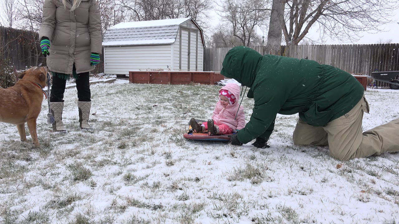 Adult kneels beside a child on a red sled in a snowy backyard.