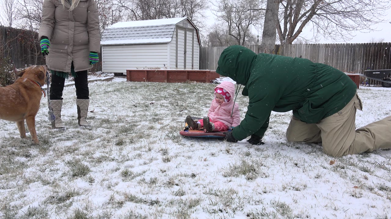 The child remains seated on the red sled in the snowy Westminster yard while the kneeling adult reaches toward the sled and the standing adult is partly visible behind them.