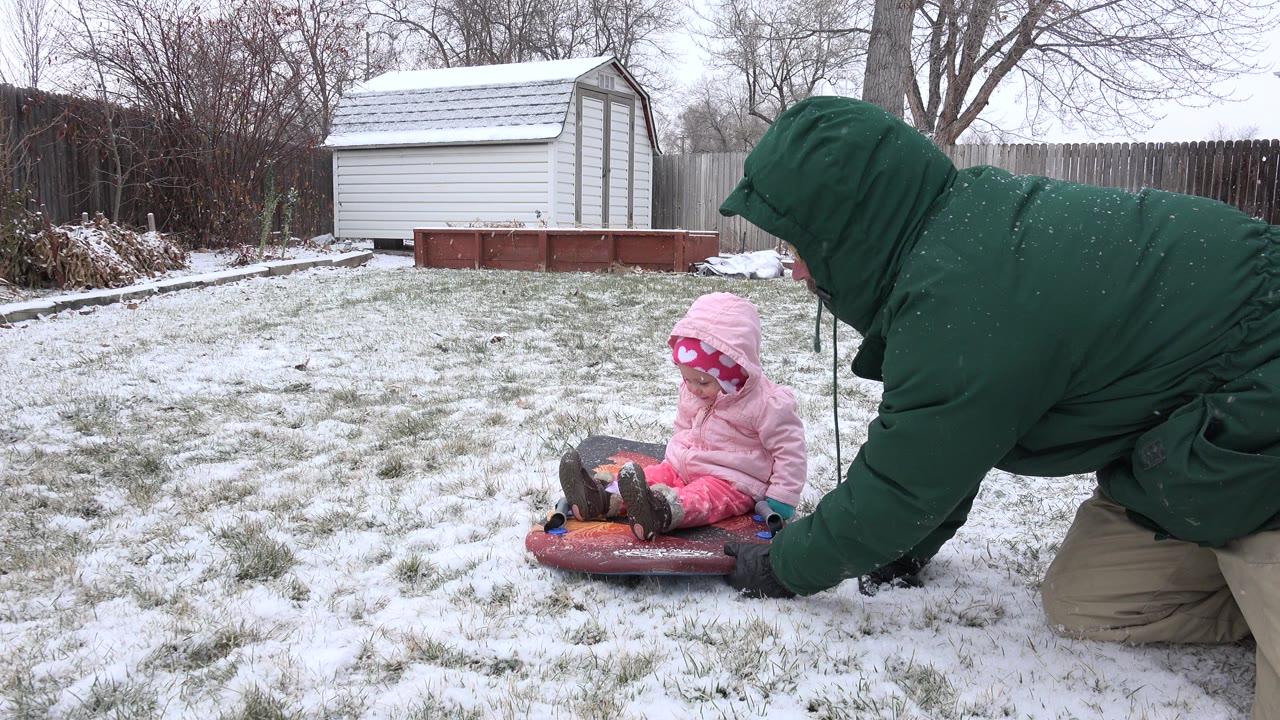 Baby in pink winter clothes sits on a red sled while an adult kneels beside it in a snowy yard.