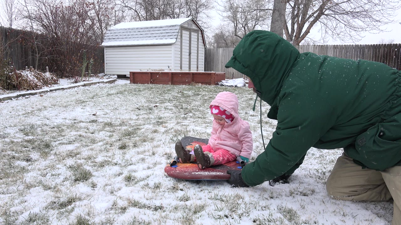In Westminster, Colorado, the adult stays low beside the sled while the baby looks downward, with snow-speckled grass, a white shed, and garden beds behind them.