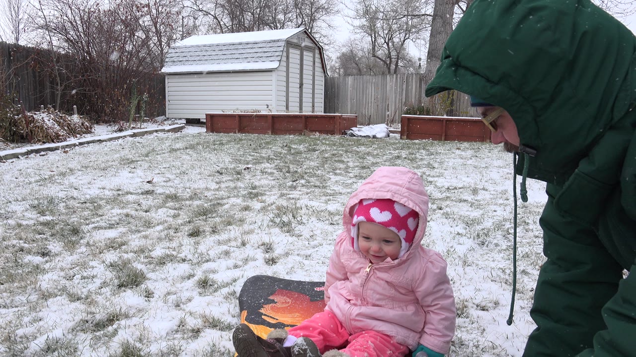 Baby in pink winter clothes sits in snowy yard beside a sled as a parent leans nearby.
