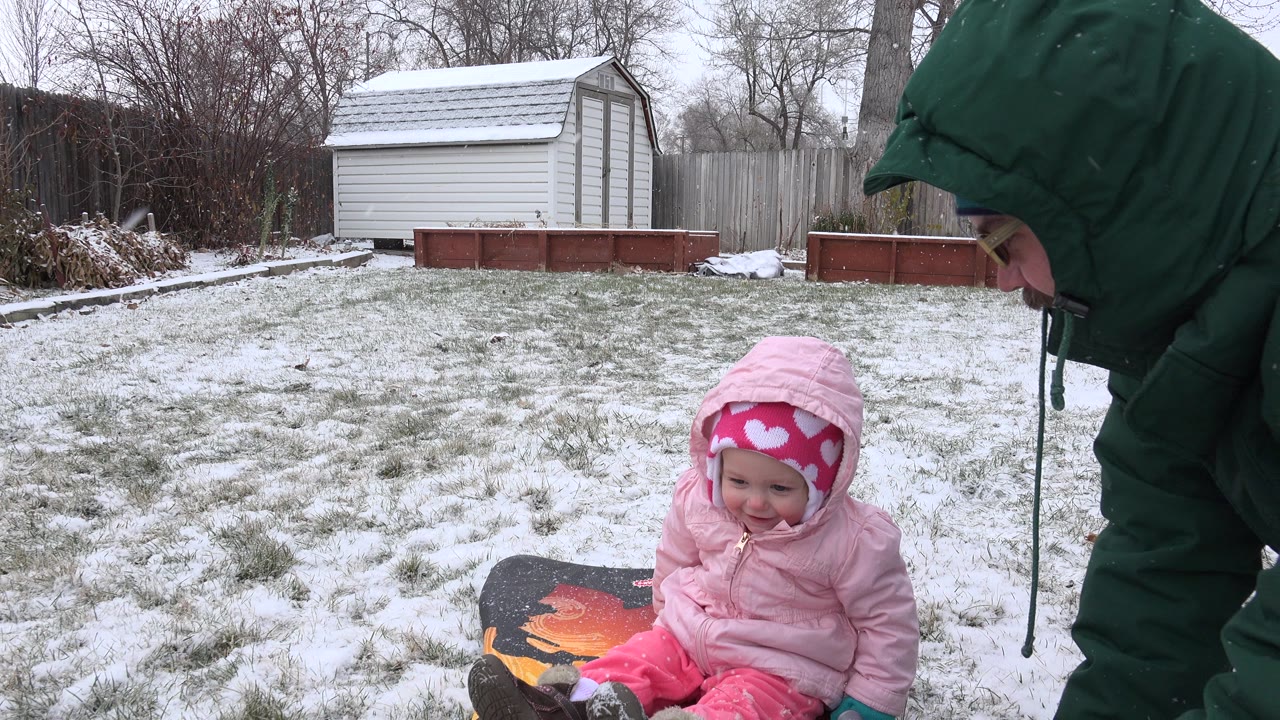 At 0:01 in Westminster, Colorado, the baby remains seated beside the sled while the parent's green coat fills the right side and the shed and fence stay visible behind the snowy lawn.