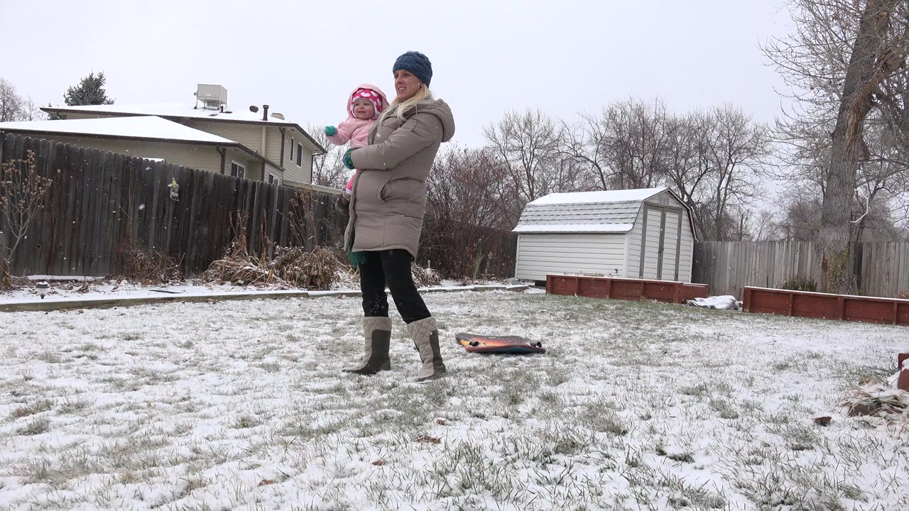 Mother holding a baby in a snowy backyard near a shed.
