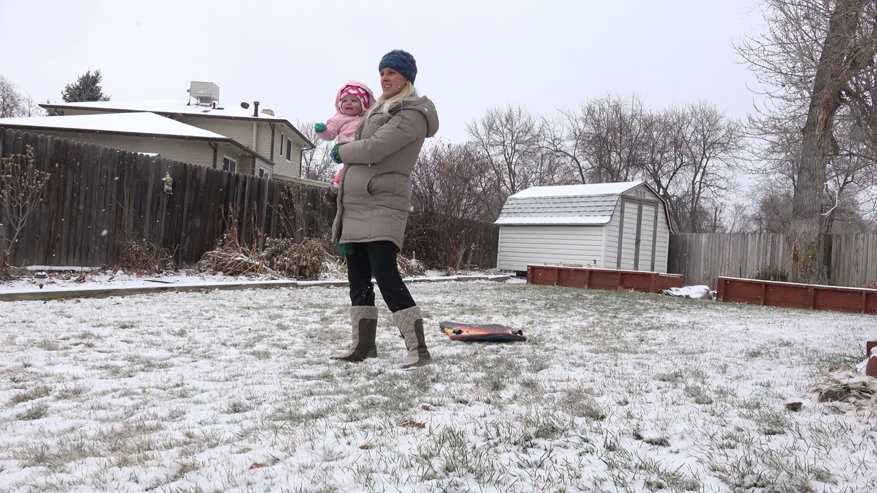 The mother and baby remain in the snowy Westminster backyard, with the baby facing outward and a small toy lying on the grass near the shed and raised garden beds.