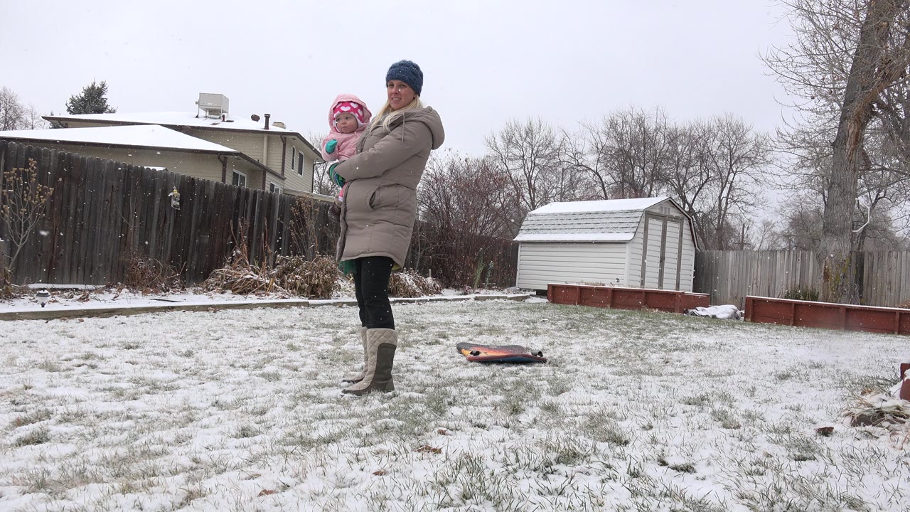 The woman and child remain in the snowy Westminster, Colorado yard, standing near the left side of frame with a small shed and fenced residential backdrop visible.