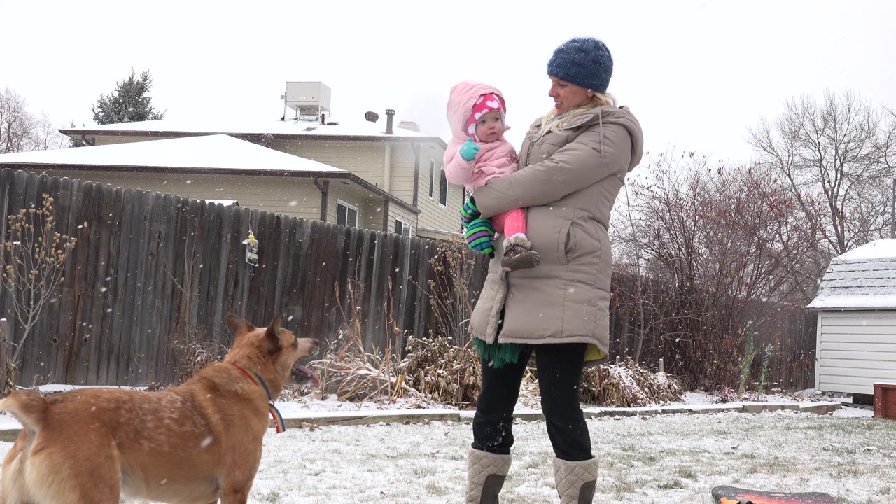 In Westminster, Colorado, a woman in winter clothing holds a small child in pink while a brown dog stands in the snowy backyard foreground.