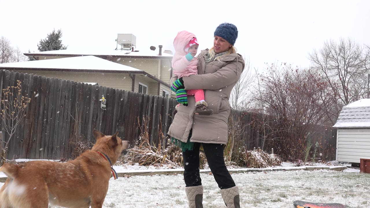 Woman holding a child turns slightly as a dog stands nearby in snow.
