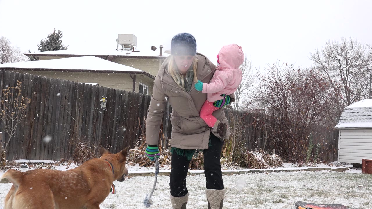 Mother holding a baby in snow while a brown dog stands in the foreground.