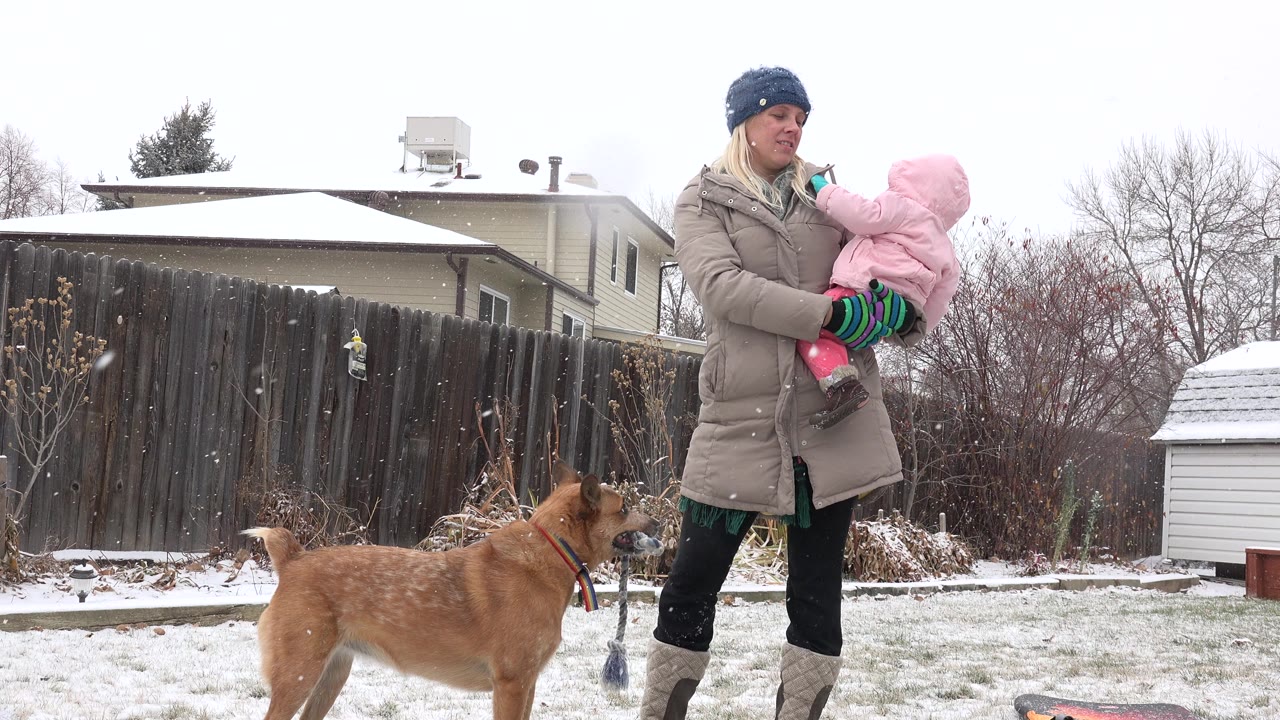 Woman holding a child in a snowy yard while a brown dog stands nearby.