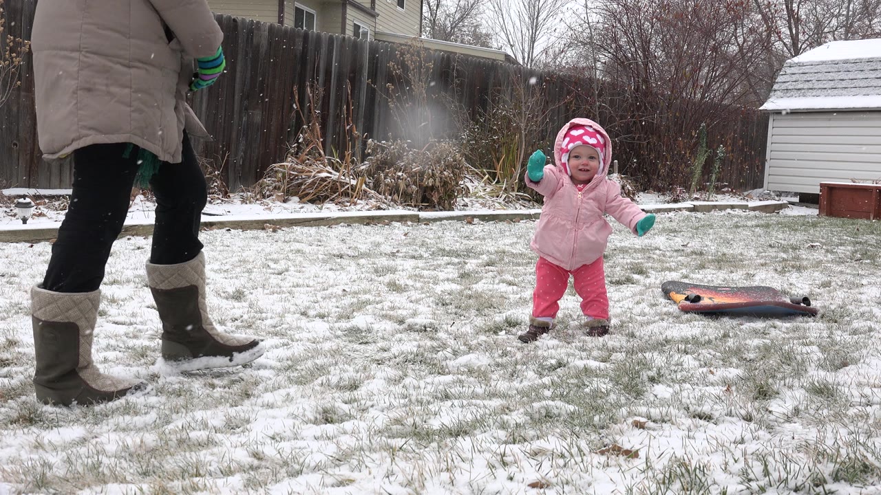 Baby in pink winter clothes stands on snowy backyard grass as an adult walks nearby.