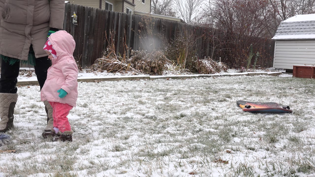 Toddler in pink snow clothes standing beside an adult on a snowy lawn.