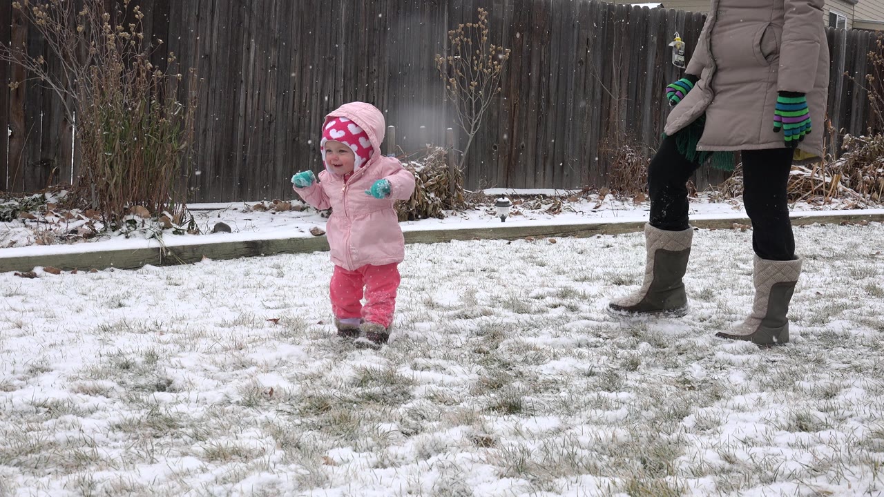 A toddler in pink snow pants, jacket, and hat walks across a lightly snow-covered Westminster, Colorado backyard while an adult woman stands partly in frame beside her.
