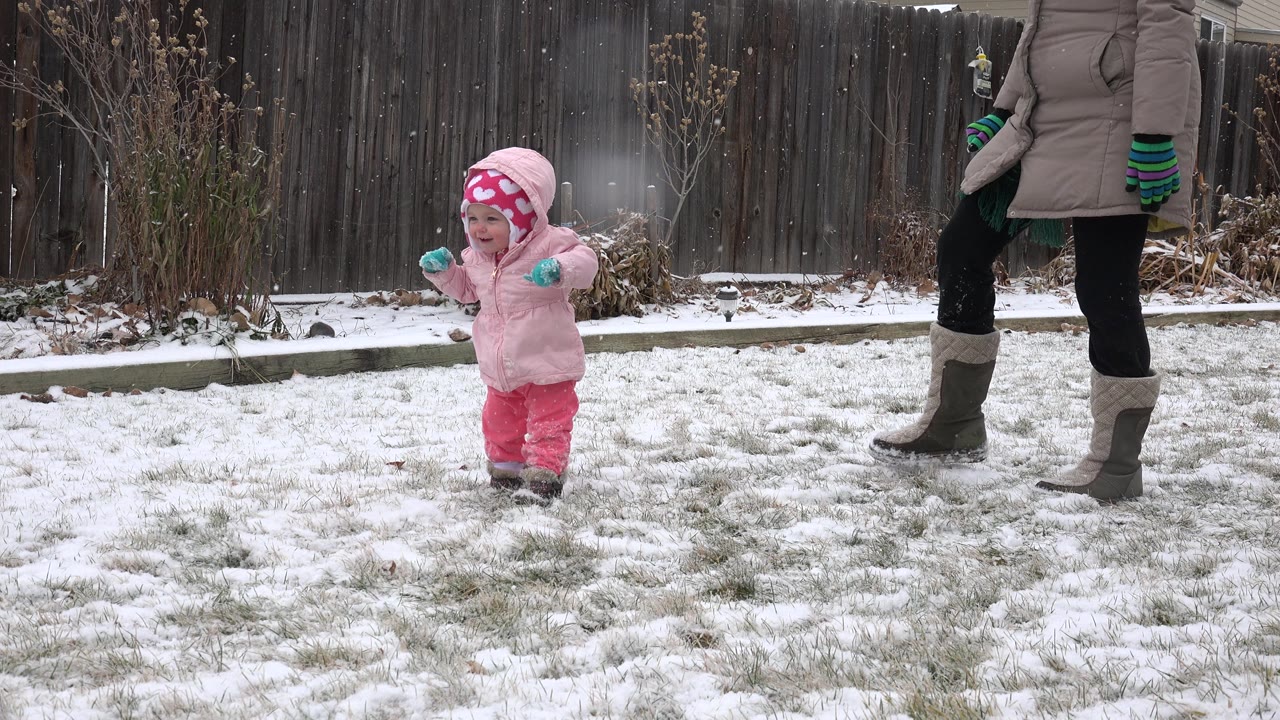 The toddler continues across the thin snow in a Westminster, Colorado backyard, with the adult woman's coat, gloves, leggings, and boots visible at the right edge.